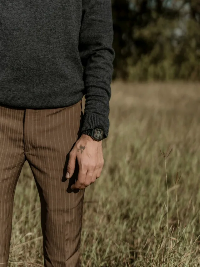 A man's hand with tattoos wearing a wristwatch stands in a grassland.