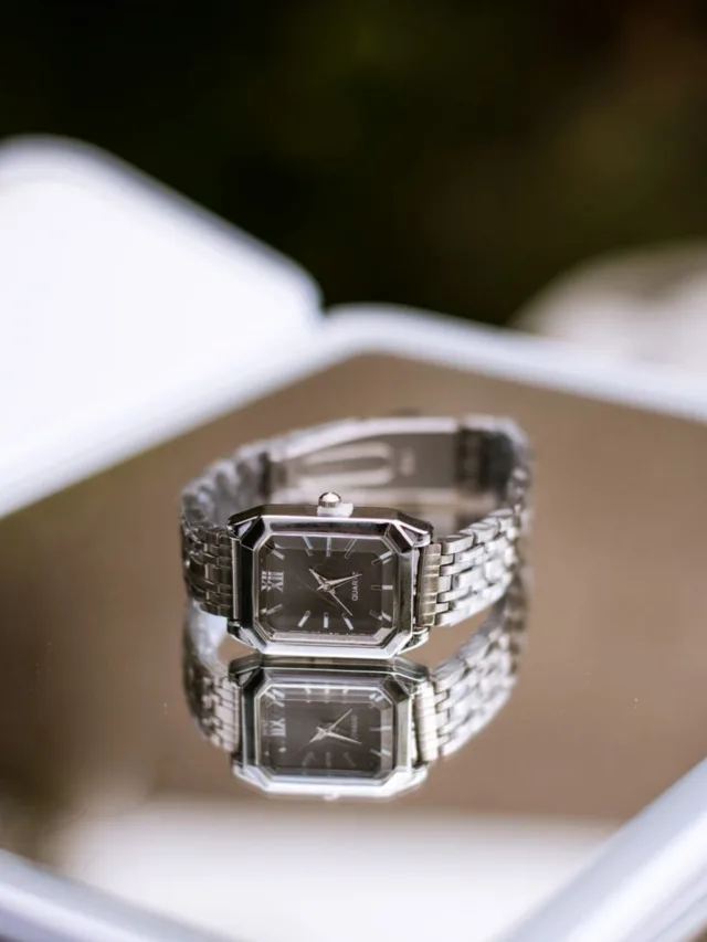 Sophisticated silver watch with black dial reflecting on a mirrored surface, showcasing its luxurious design.