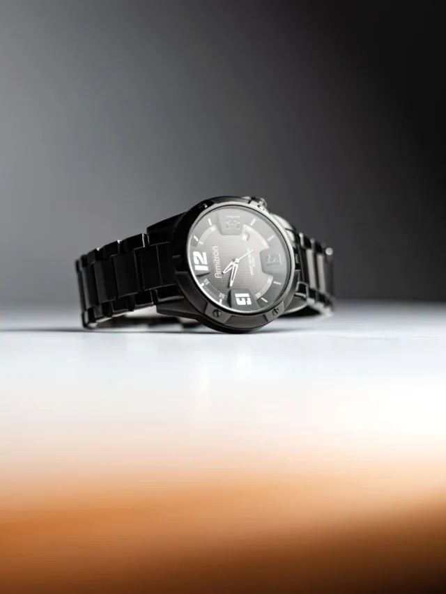 Elegant black wristwatch with a glossy finish, displayed on a reflective surface with a soft background blur.